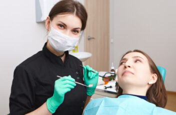 Indoor shot of skillful confident young female dentist wearing exam gloves and white mask holding metal probe and dental miror, ready to examine oral cavity of woman patient sitting in chair