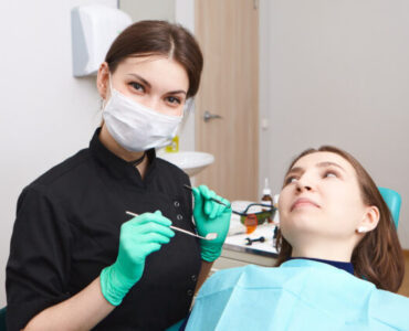 Indoor shot of skillful confident young female dentist wearing exam gloves and white mask holding metal probe and dental miror, ready to examine oral cavity of woman patient sitting in chair